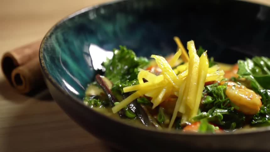 A ceramic bowl is filled with a shrimp curry as additional sliced mango is dropped on top of it