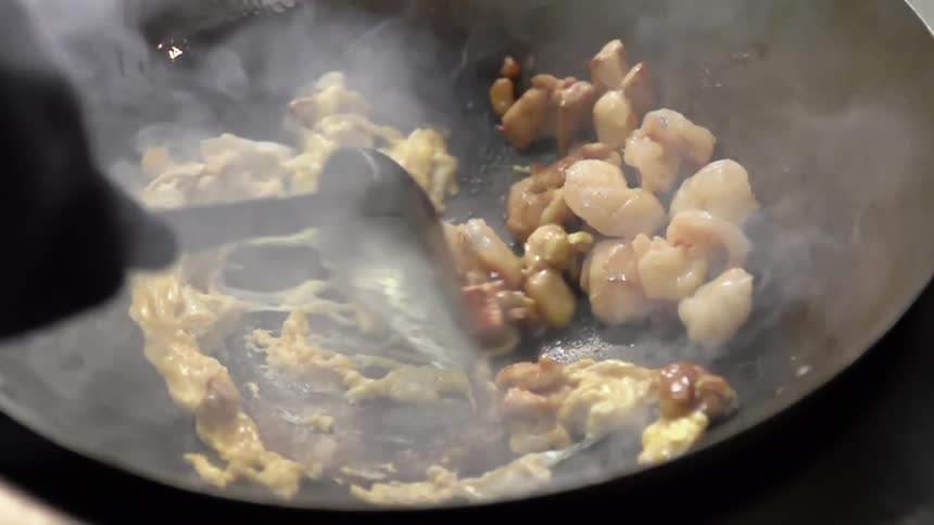 A gloved hand stirs eggs in a steaming smoking wok with shrimp and vegetables