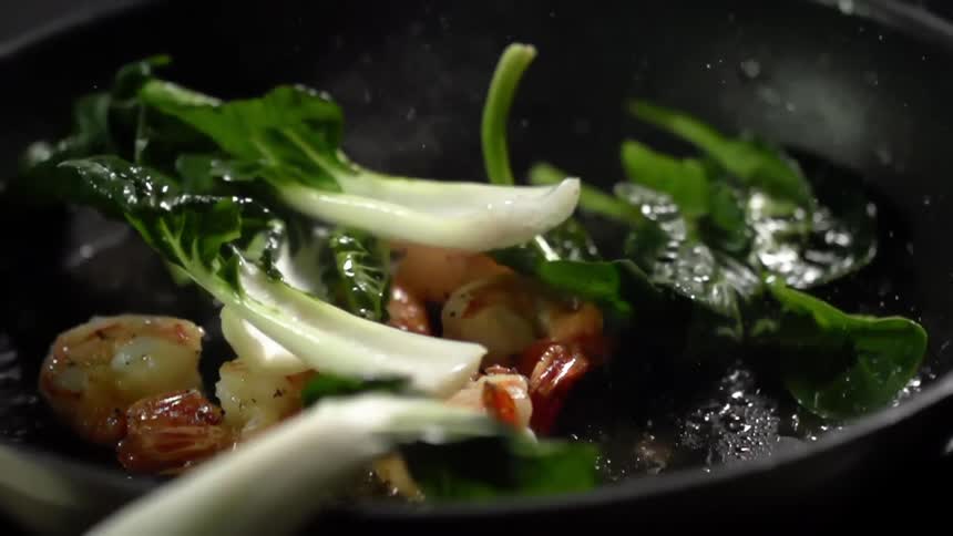 Juicy shrimps are being roasted while bok choy is being dropped into the black skillet