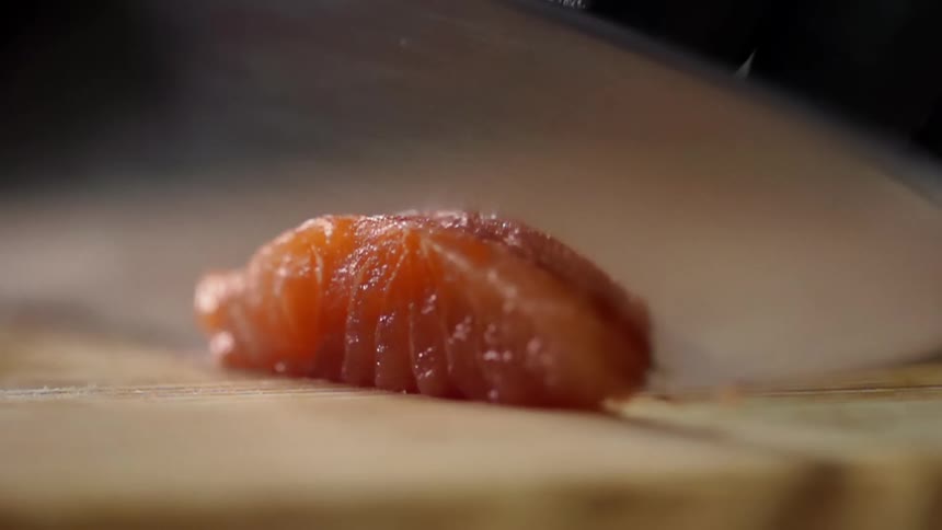 A person uses a large kitchen knife to skillfully slice a thin piece of raw salmon