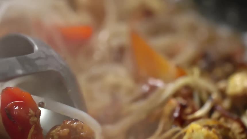 A spoon stirs a noodle and vegetable meal in a pan