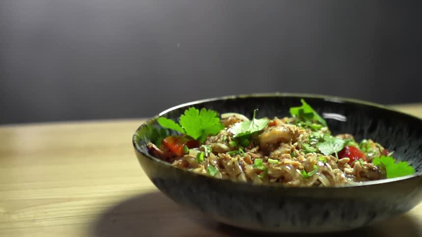 A bowl of noodles on a table with leafy vegetables falling into the bowl