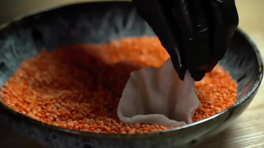 Chef wearing black gloves places rice chips over red lentils in a black bowl
