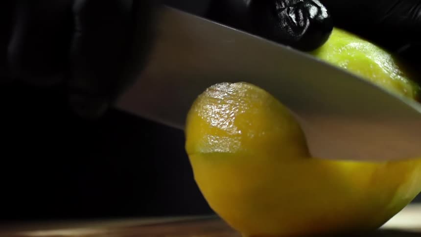 One hand covered with a black glove secures a mango as the other holds a chefs knife and makes a thin slice