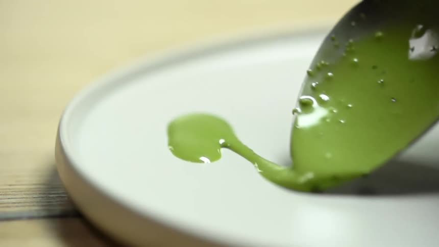 A small spoon is used to drizzle a bright green sauce onto a glass plate