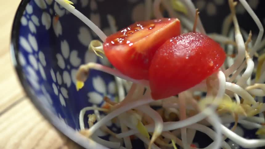 Sliced tomato falls over bean sprouts inside a blue ceramic bowl over a wooden table
