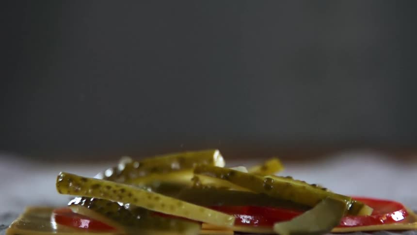 Thin pickle wedges drop onto tomato slices against grey background