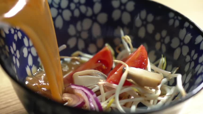Soup broth is poured into a small glass bowl that is filled with a variety of vegetables like tomatoes and bean sprouts