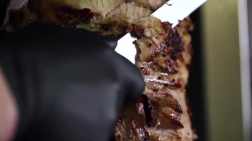 Grilled meat on a vertical spit is carefully carved with a knife by a person wearing dark rubber gloves