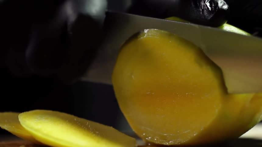 Cook with black gloves slices a mango thinly on a cutting board in studio