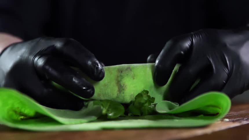 A cook wearing black gloves wraps vegetables in a green tortilla on a wooden table