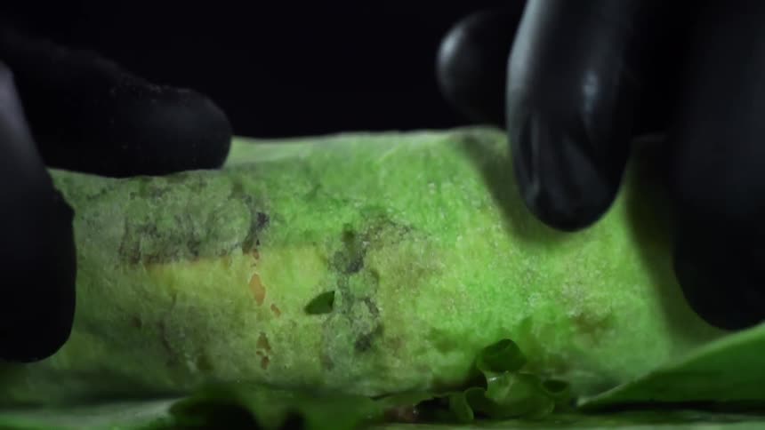 A pair of hands covered in black gloves wrap vegetables in a green tortilla