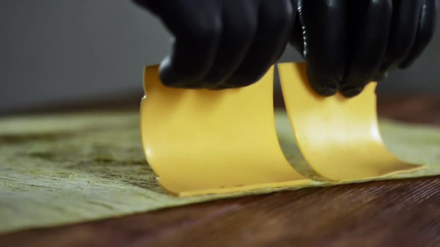 Two hands wearing black gloves place a slice of cheddar cheese on a tortilla