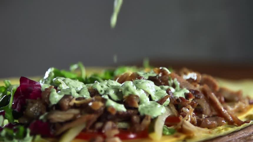 A greenish sauce pours onto meat and vegetables lying on a green tortilla