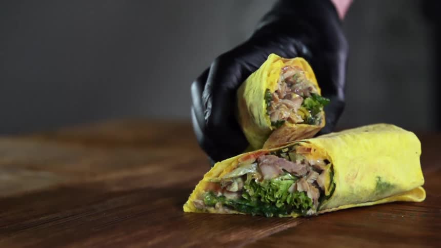 A gloved hand places a sliced wrap sandwich half on top of the other half on a wood table