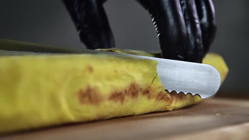 A gloved hand holds down a wrap as a serrated knife cuts through with sauce sticking to the blade