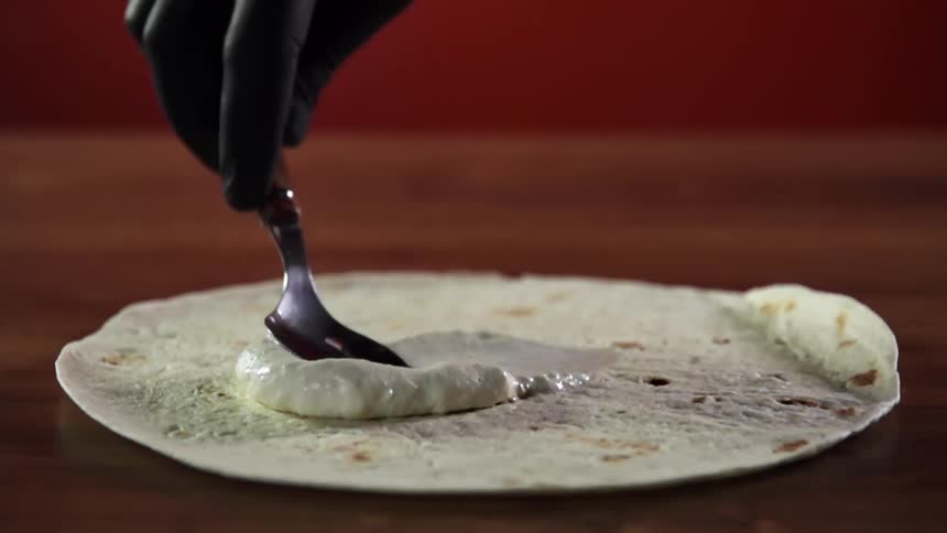 A person with black gloves holding a spoon spreading a paste on flatbread
