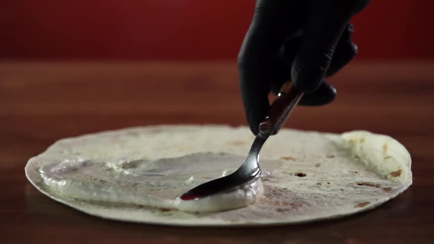 Chef wearing black leather gloves spreads white sauce over a flour tortilla using a metallic spoon