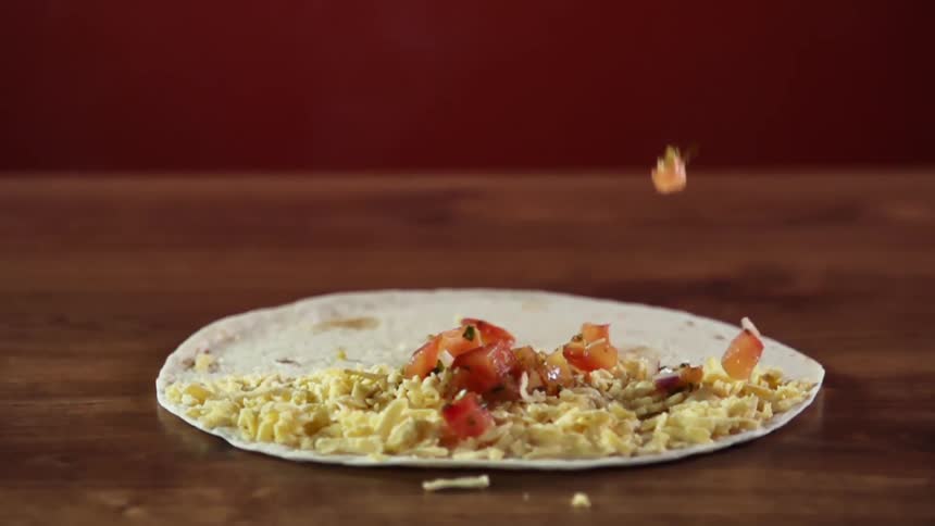 Diced tomatoes falling onto shredded cheese on a tortilla lying on a wooden table