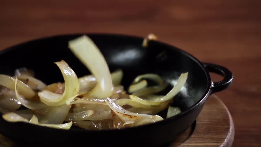 Slices of cooked onions drop inside a black cast iron pan over a wooden table