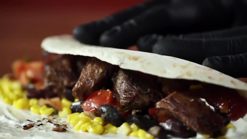 Chef wearing black latex gloves wraps a flour tortilla over meat and vegetables