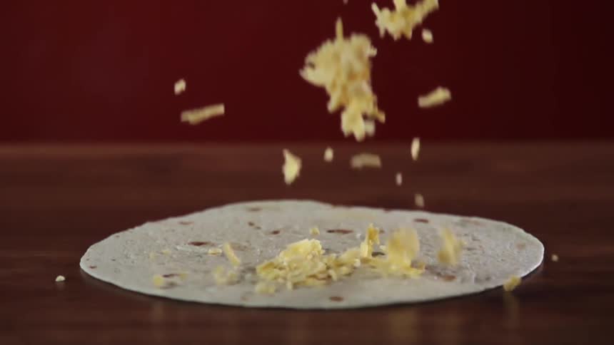 Shredded cheese being poured on a tortilla lying on a wooden table