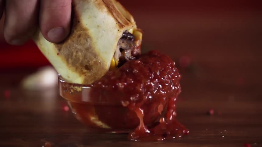A man dunks a quesadilla into a small bowl of salsa and sends salsa flying and spilling on the table
