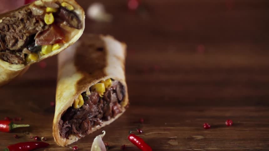 A person lifts a beef wrap from atop another wrap on a wooden table