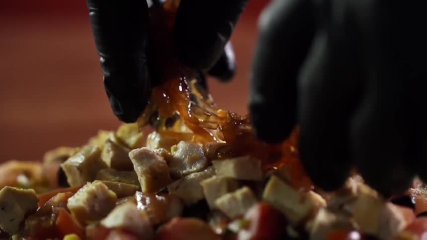 A person wearing black gloves squeezes sticky sauce over a pile of food