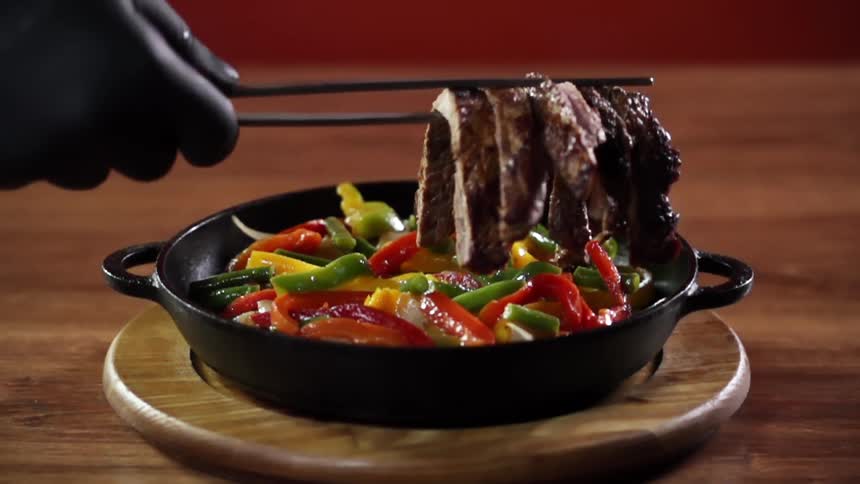 A gloved hand places strips of beef in a small skillet filled with sliced peppers using a pair of food tweezers