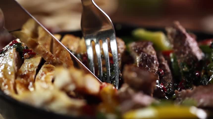 A knife and a fork dig into a meal in a black bowl