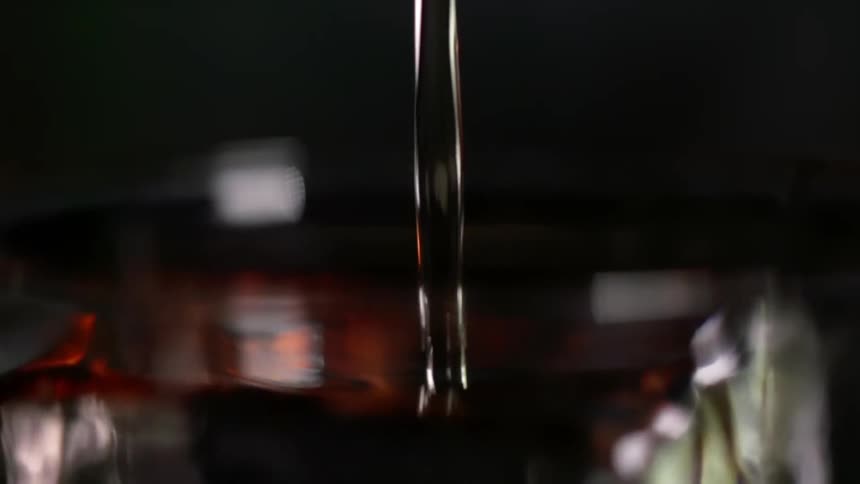 Cocktail is being prepared into a glass with an ice cube on dark background