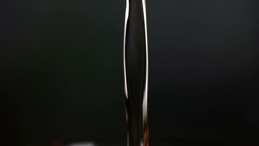 Cocktail is being poured into a glass with an ice cube on dark background