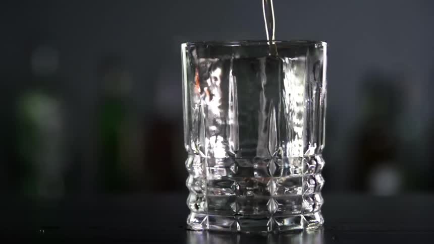 Cocktail is poured into a glass on a bar counter with bottles lined up behind it