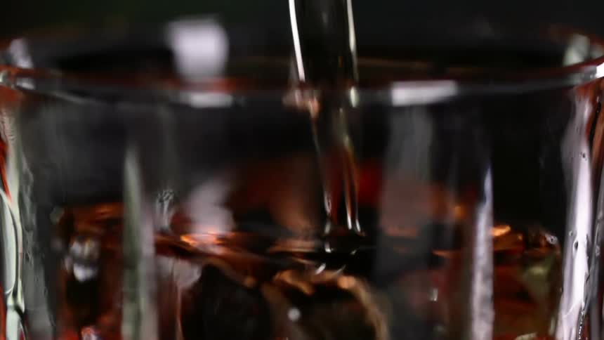 Cocktail is being poured into a glass with an ice cube on dark background