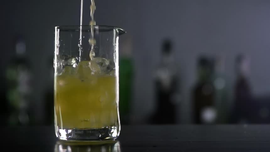 A cocktail being poured into a glass with ice cubes in a bar