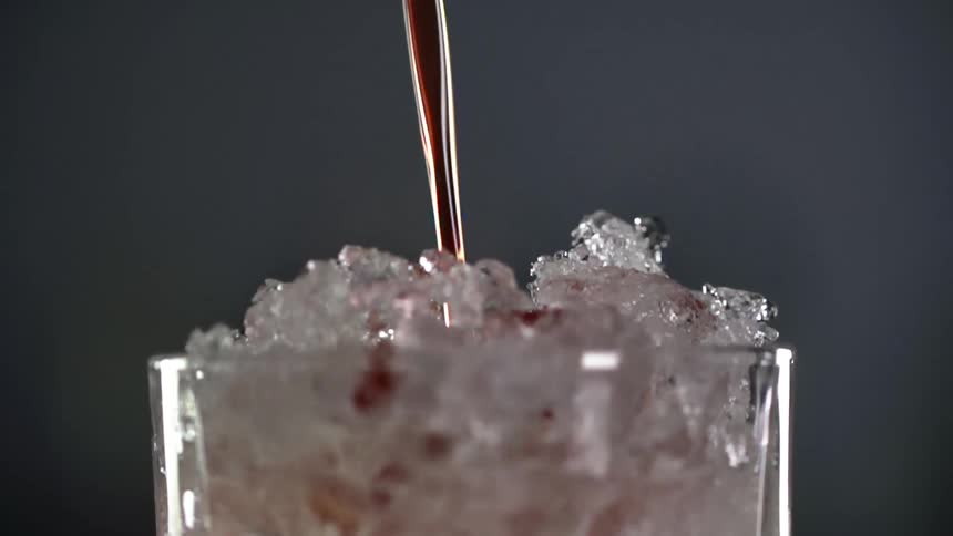An amber alcoholic beverage pours into a drinking glass filled with crushed ice