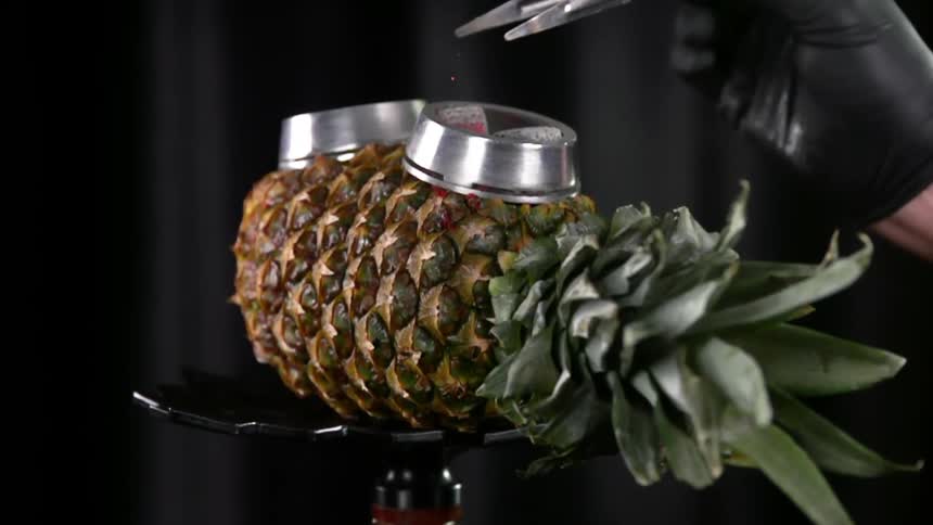 Charcoal cubes being put in a metal ring over a pineapple used as a bowl with a tweezer