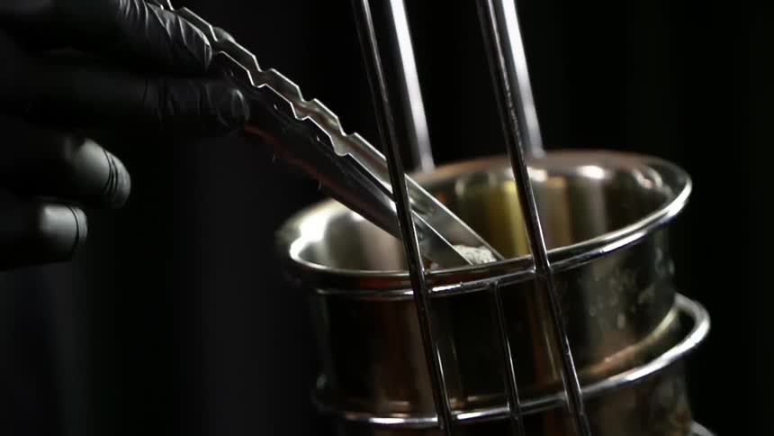 A person uses small tongs to pick a charcoal cube up out of a metal bowl