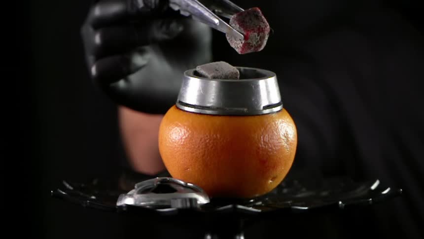 A person places coals on top of an orange used as a bowl for hookah