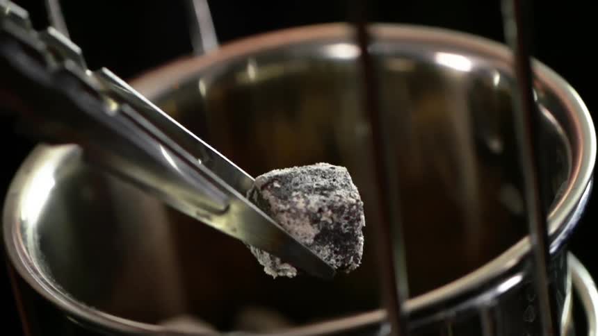 A person uses small tongs to pick a charcoal cube up out of a metal bowl
