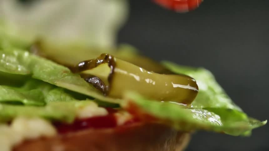 A tomato being dropped on a bun with lettuce and pickles