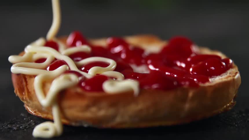 Mayonnaise is slowly drizzled atop a sliced bun that is already coated with ketchup