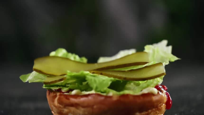 Two thinly sliced pickles land upon a bed of lettuce that sits on the bottom of a brioche bun as ketchup drips off the side