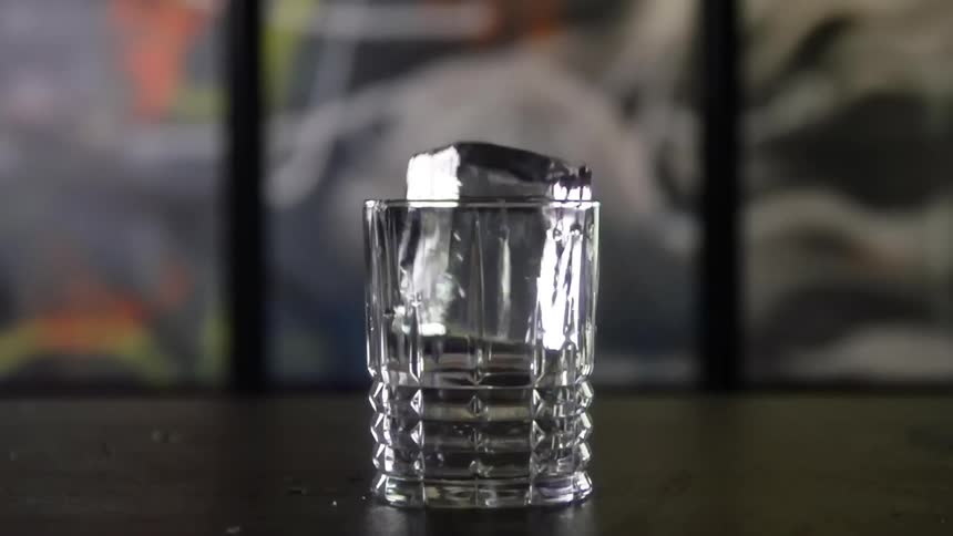 A shot glass on a table with a ice cube falling into it