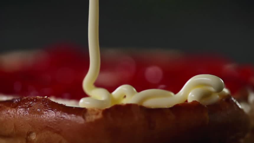 Close up of a mayonnaise that is slowly drizzled atop a sliced bun that is already coated with ketchup