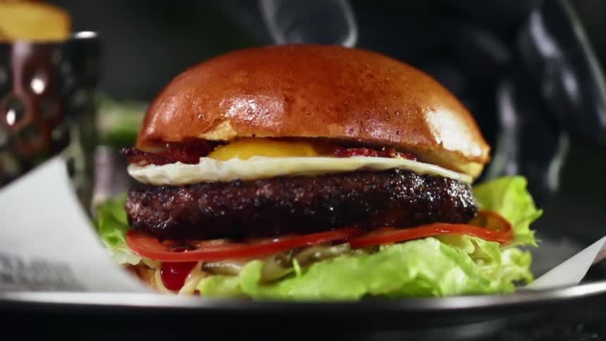 Hand wearing black glove gently places a hamburger on a brioche bun  on top of parchment paper that is placed on a plate
