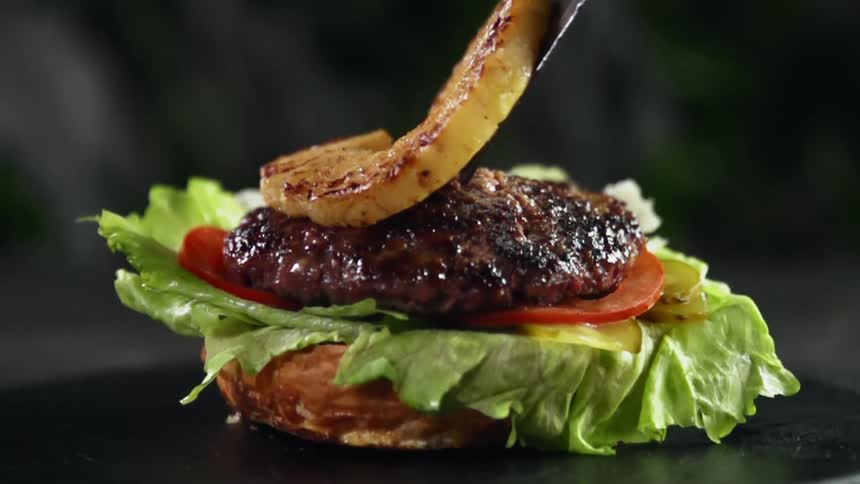 A spatula drops a piece of grilled pineapple on top of a burger patty