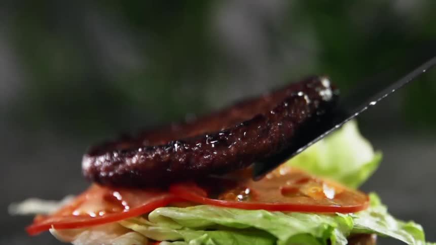 A burger patty being placed on lettuce and tomatoes