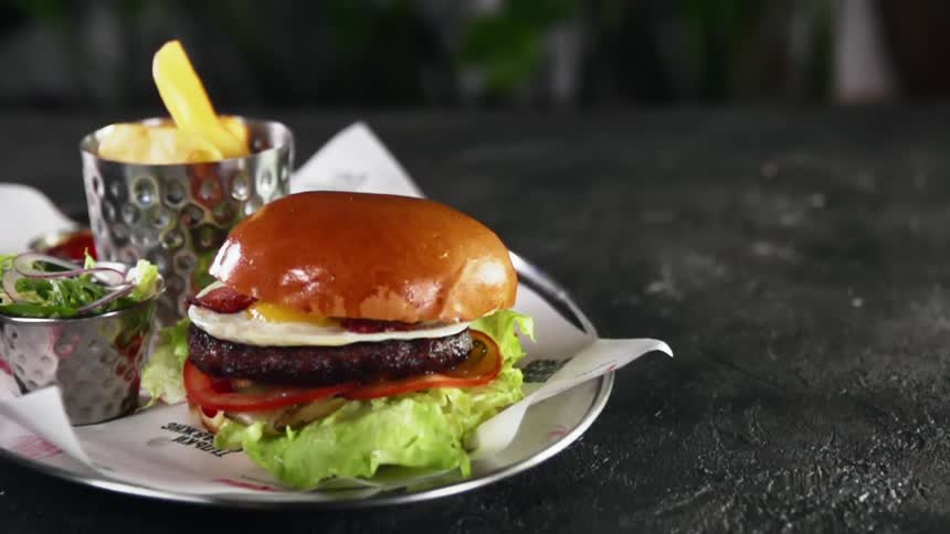 A hamburger on a plate with the bun falling on top of the patty with fried egg and bacon
